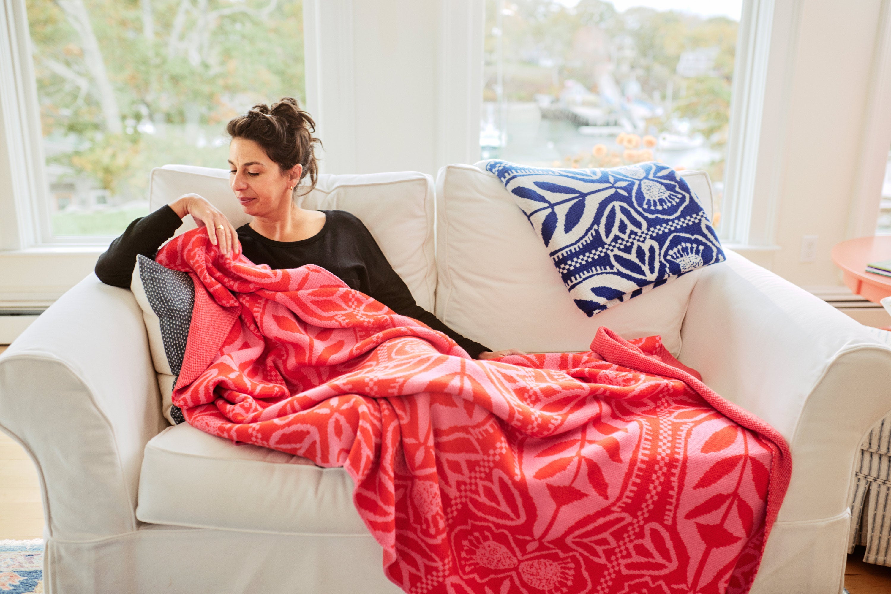 Woman sitting on a white couch with a pink patterned blanket, surrounded by large windows.